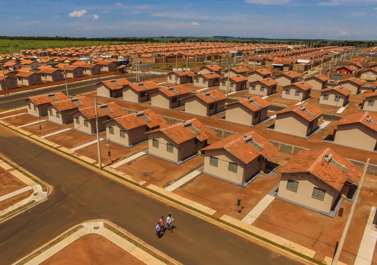 Vista aérea de um conjunto habitacional popular. Várias casas iguais desocupadas e ruas vazias.