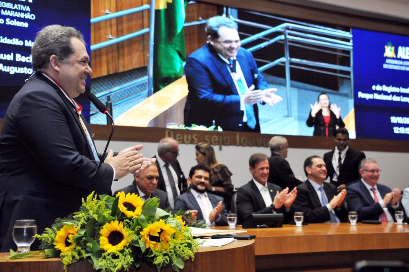 Foto horizontal da entrega de registro do Parque Nacional dos Lençóis Maranhenses. Um homem, que é o homenageado da solenidade (o ministro Carlos Augusto Pires Brandão), está em pé à esquerda, sorrindo e aplaudindo. Ele veste um terno e usa uma medalha no peito. Um microfone está posicionado à sua frente. A mesa de madeira está em primeiro plano, com um arranjo de flores amarelas (girassóis e folhagens) em destaque. Vários homens em trajes formais (ternos) estão sentados à mesa, também aplaudindo. A grande tela de vídeo no fundo exibe: Um close-up (em tempo real) do orador no púlpito. Trechos do texto sobre a solenidade, incluindo menções à "Entrega do Título de Cidadão Maranhense" e a "Medalha Manuel Beckman" ao Ministro Carlos Augusto Pires Brandão. É possível ver uma intérprete de Libras no canto inferior direito da projeção.