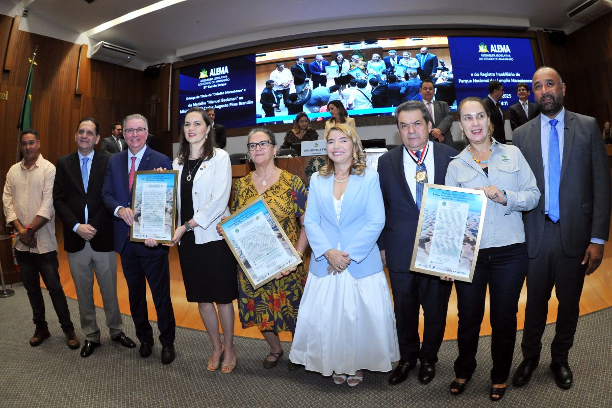 Foto horizontal da entrega de registro do Parque Nacional dos Lençóis Maranhenses. Há um grupo de oito pessoas posando para uma foto no centro da imagem. Elas estão vestidas de maneira formal ou semi-formal. Várias pessoas no grupo central estão segurando quadros emoldurados de registros. Uma grande tela de vídeo no fundo exibe a logomarca da ALEMA (Assembleia Legislativa do Estado do Maranhão) e um texto que menciona a "entrega do registro imobiliário do Parque Nacional dos Lençóis Maranhenses". Há a presença de uma bandeira do Maranhão e a iluminação típica de um evento interno. Há reflexos e imagens de outras pessoas no fundo, tanto na tela de vídeo quanto na sala, indicando a presença de uma audiência ou convidados.