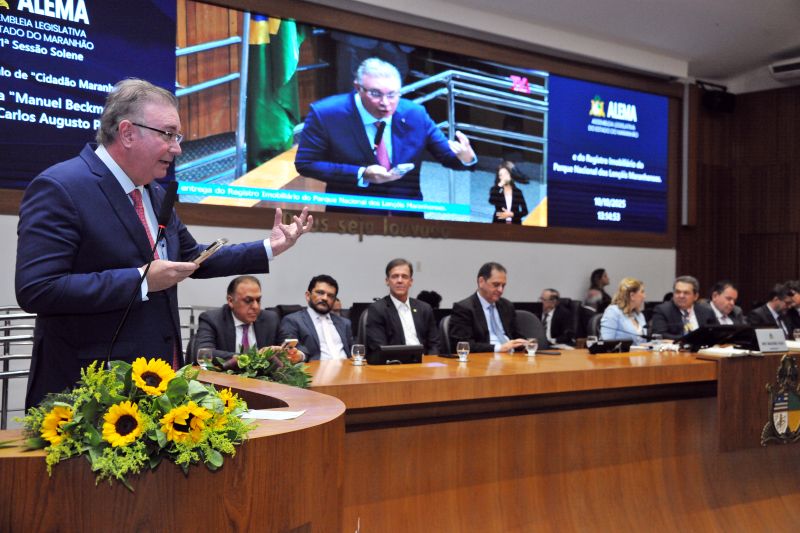 Foto horizontal da entrega do registro do Parque Nacional dos Lençóis Maranhenses. O desembargador Froz Sobrinho está em pé, à esquerda, fazendo um discurso. Ele veste um terno escuro, segura um objeto pequeno na mão esquerda e gesticula com a mão direita, com um microfone à sua frente. Diversas autoridades, todas de terno, estão sentadas à mesa, atentas ao orador. Um arranjo de flores amarelas (girassóis) decora a mesa em primeiro plano. A grande tela de vídeo exibe informações sobre o evento: À esquerda, os detalhes da homenagem. 