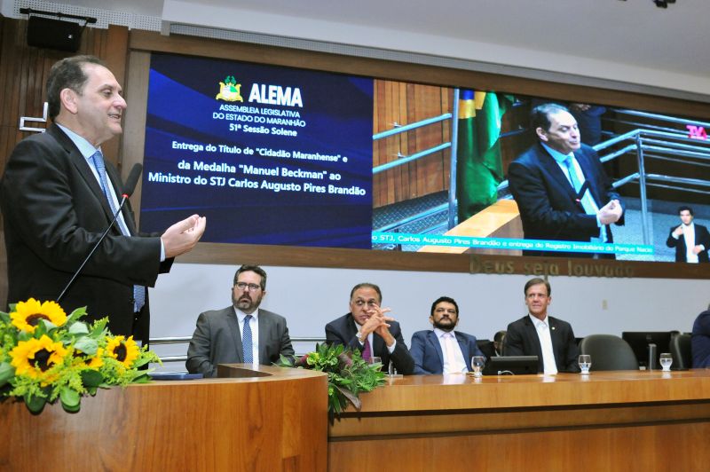 Foto horizontal da entrega do registro do Parque Nacional dos Lençóis Maranhenses. O ministro Reynaldo Soares da Fonseca está em pé à esquerda, discursando. Ele veste um terno escuro com uma gravata azul-clara e gesticula com as mãos. Em primeiro plano, uma parte da mesa de madeira está decorada com um arranjo de girassóis. Quatro autoridades estão sentadas, atentas ao orador. A grande tela de vídeo no fundo fornece o contexto do evento: Exibe a logomarca da ALEMA.